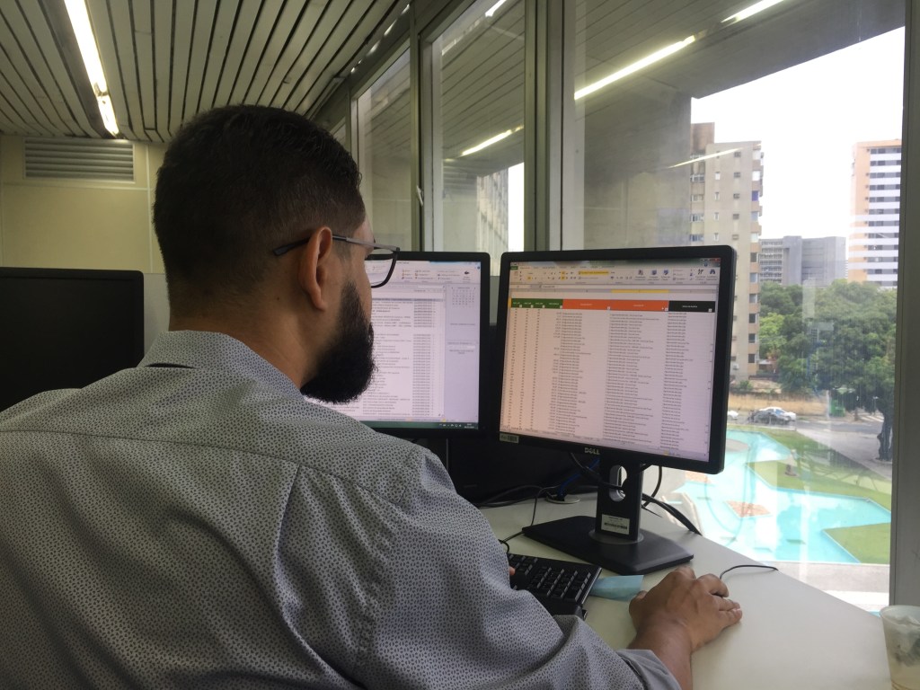 A person work in a computer with 2 screens. A window and the view of the street in background.
